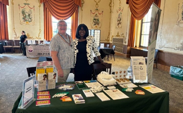 LaShawnda Bates, wearing a black dress with a white polka-dot blazer, and Andrew Lowden, wearing a striped button-up shirt with rainbow lanyard, stand together in front of an informational booth at a health fair.